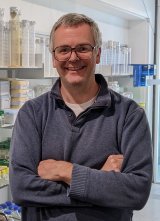 Photograph of Colin in a navy sweatshirt, with a laboratory in the background.